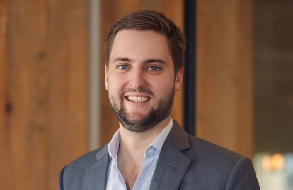 A person wearing a grey suit jacket over a light blue button-up shirt stands indoors near a wooden wall and glass partition.