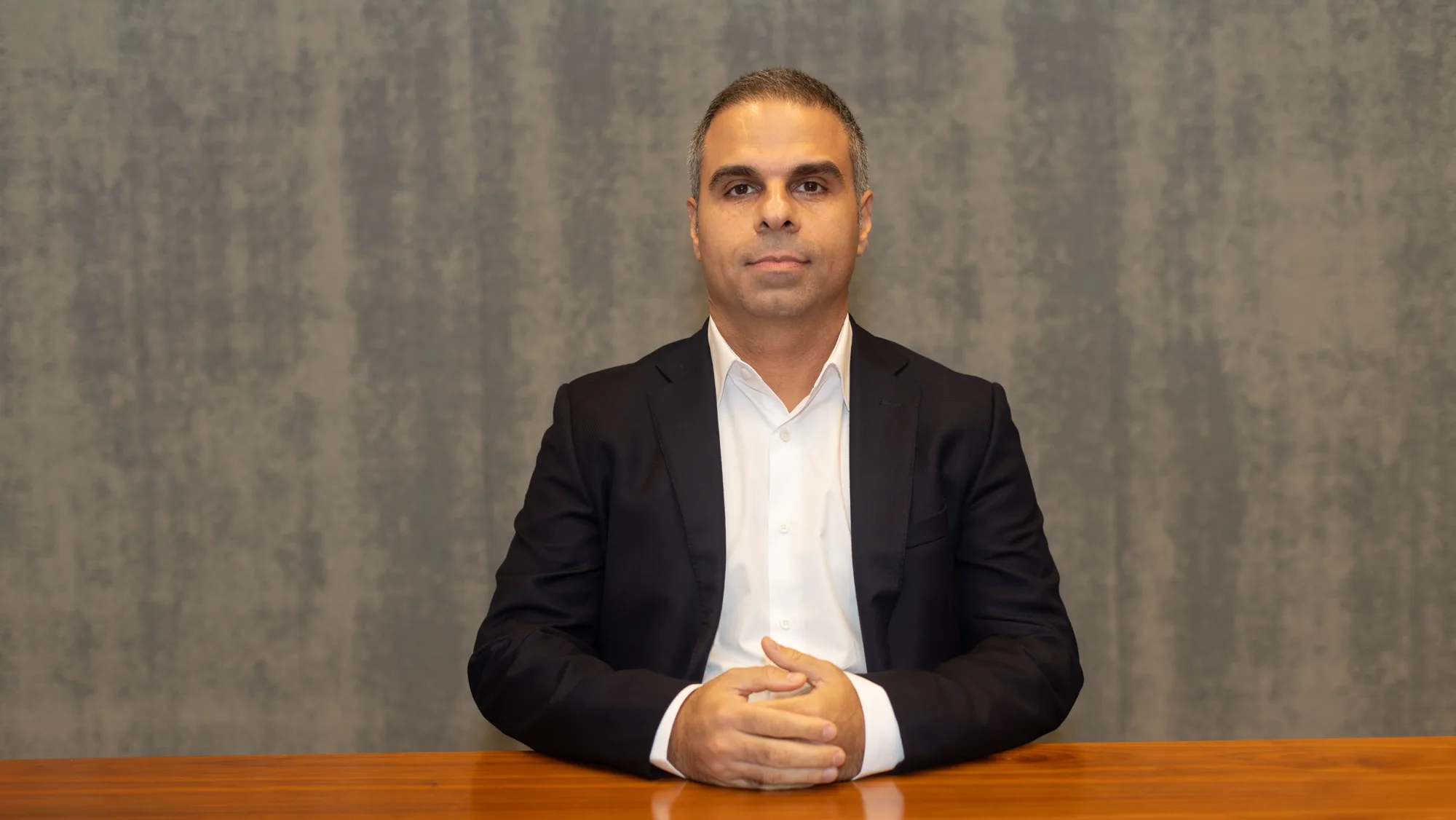 Fazel in a dark suit and white shirt sits at a wooden table with hands clasped. The background features a textured grey wall.