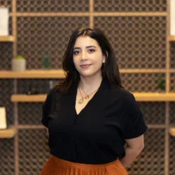 A person with shoulder-length dark hair wearing a black top and orange skirt stands in front of a patterned wall with wooden shelves holding framed certificates, geometric sculptures, and potted plants.