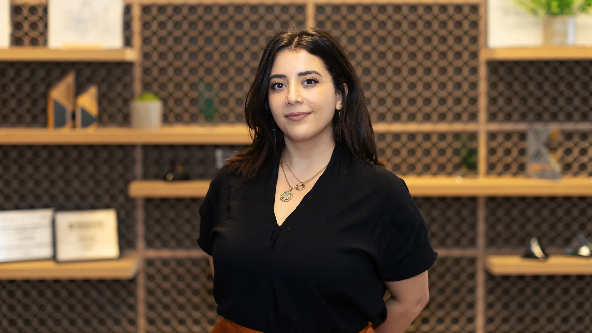 A person with shoulder-length dark hair wearing a black top and orange skirt stands in front of a patterned wall with wooden shelves holding framed certificates, geometric sculptures, and potted plants.