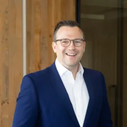 Person wearing a blue suit and white shirt stands with hands in pockets in an office with wooden paneling, glass walls, and ceiling fixtures visible in the background.