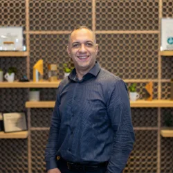 A person in a blue striped shirt stands before a wall with hexagonal patterns and wooden shelves holding framed certificates, small potted plants, awards, and decorative objects.