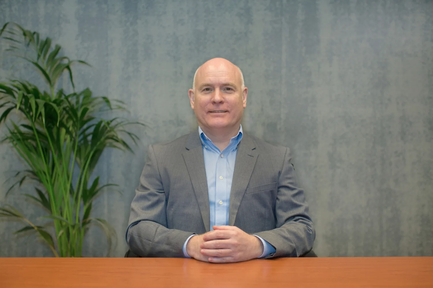 Richard wearing a pale blue shirt and grey suit jacket sat at a wooden table in front of a plant and a grey wall