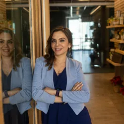 A person in a blue dress and light gray blazer stands indoors with arms crossed, leaning against a glass wall that reflects their figure. The hallway behind them is lined with shelves, plants, and framed items.