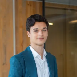 Portrait of a person with short dark hair wearing a teal blazer and white button-up shirt, standing indoors near glass walls and vertical wooden panels, with office lighting visible in the background.