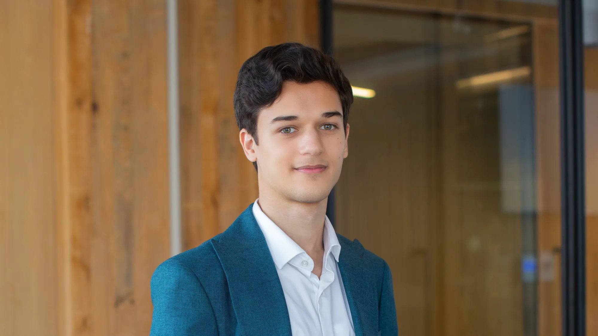 Portrait of a person with short dark hair wearing a teal blazer and white button-up shirt, standing indoors near glass walls and vertical wooden panels, with office lighting visible in the background.