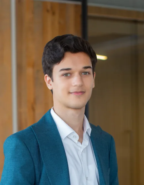 Portrait of a person with short dark hair wearing a teal blazer and white button-up shirt, standing indoors near glass walls and vertical wooden panels, with office lighting visible in the background.