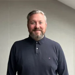Person wearing a dark long-sleeve polo shirt stands indoors against a grey wall. The room features slanted ceilings and a skylight window on the right side.