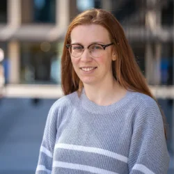 Holly in front of the office exterior wearing a grey jumper with white horizontal stripes.
