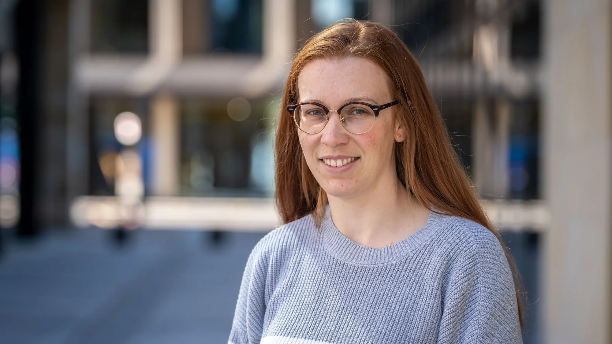Holly in front of the office exterior wearing a grey jumper with white horizontal stripes.
