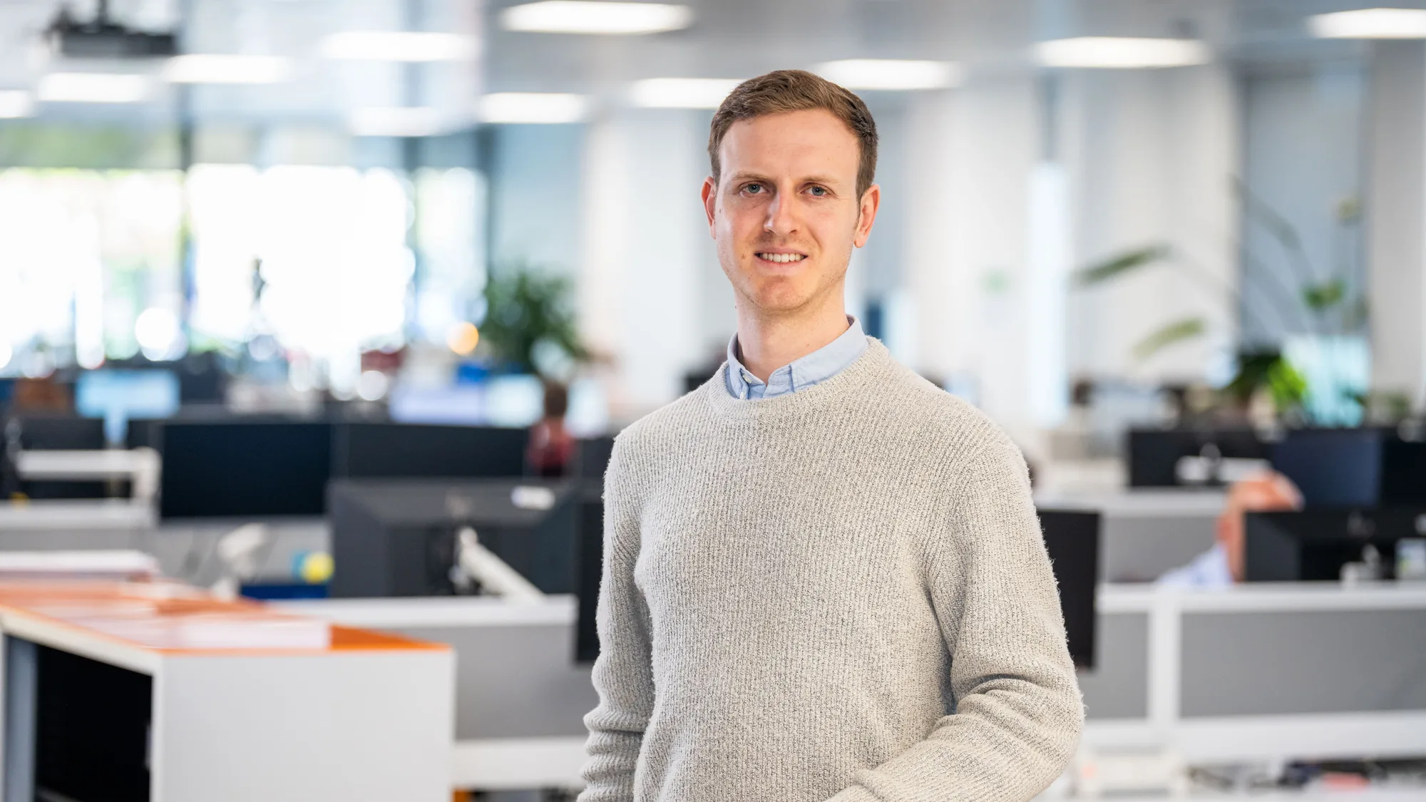Hugh in a pale blue shirt and beige jumper standing in a well lit office