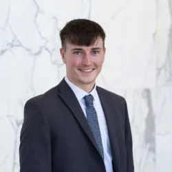 Jude stands in front of a light marble wall, wearing a dark textured suit jacket, white shirt, and patterned blue tie. Upper body portrait with neutral lighting and a clean background.