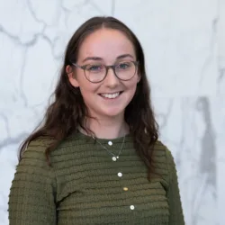Person wearing an olive-green textured long-sleeve top with white and yellow buttons and a silver necklace with pendant, standing against a light grey marble wall with darker veining.