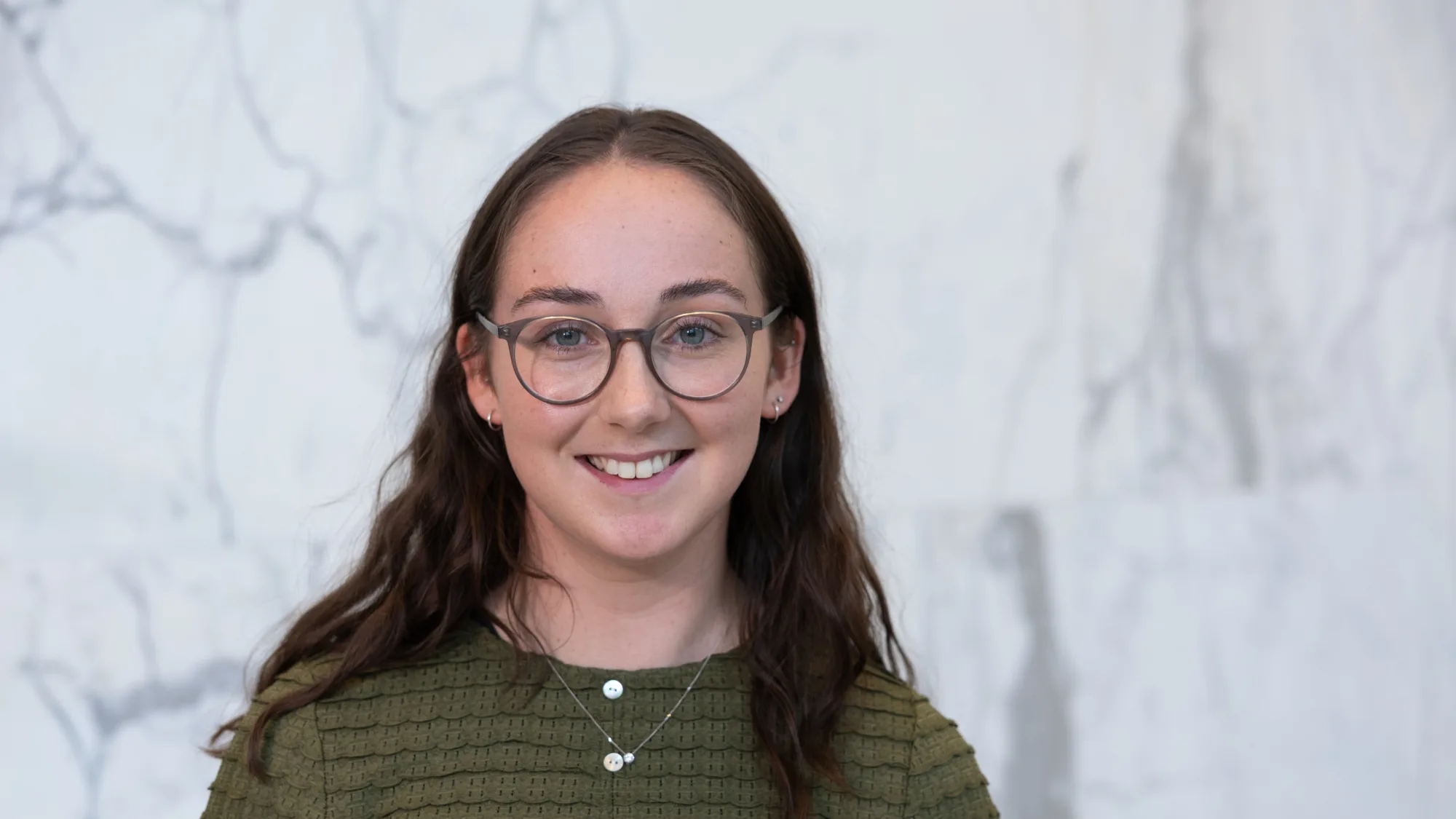 Person wearing an olive-green textured long-sleeve top with white and yellow buttons and a silver necklace with pendant, standing against a light grey marble wall with darker veining.