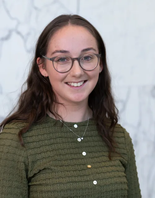 Person wearing an olive-green textured long-sleeve top with white and yellow buttons and a silver necklace with pendant, standing against a light grey marble wall with darker veining.