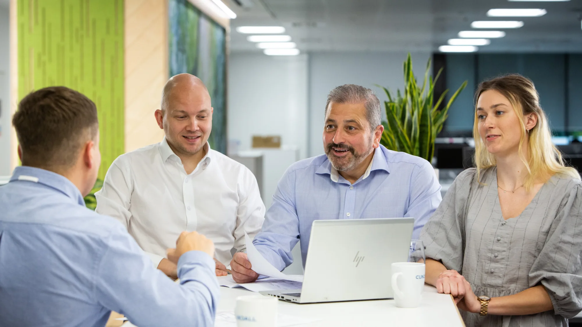 Tony Centola in Edinburgh office team meeting with laptop and mugs on the table. The background in the main office area and is blurred.
