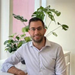 A person in a light striped shirt sits at a white desk near tall windows. Several green potted plants are arranged behind them, with a large-leaf plant against a neutral wall.
