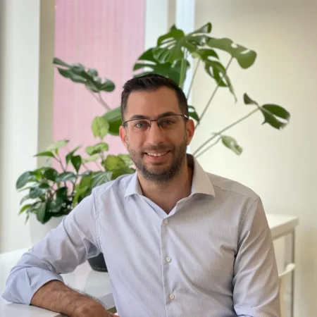 A person in a light striped shirt sits at a white desk near tall windows. Several green potted plants are arranged behind them, with a large-leaf plant against a neutral wall.
