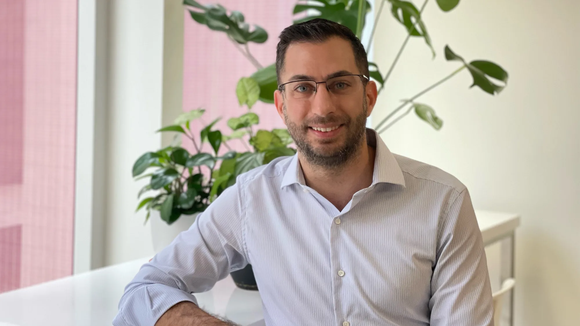 A person in a light striped shirt sits at a white desk near tall windows. Several green potted plants are arranged behind them, with a large-leaf plant against a neutral wall.