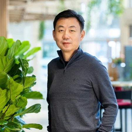 Dong stood in an open plan office with hands in pockets next to a tall green plant.