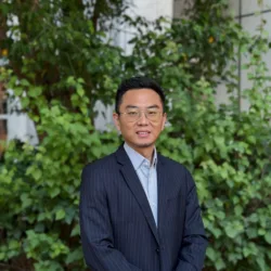 Kevin in a light blue shirt and dark suit in front of a leafy green border