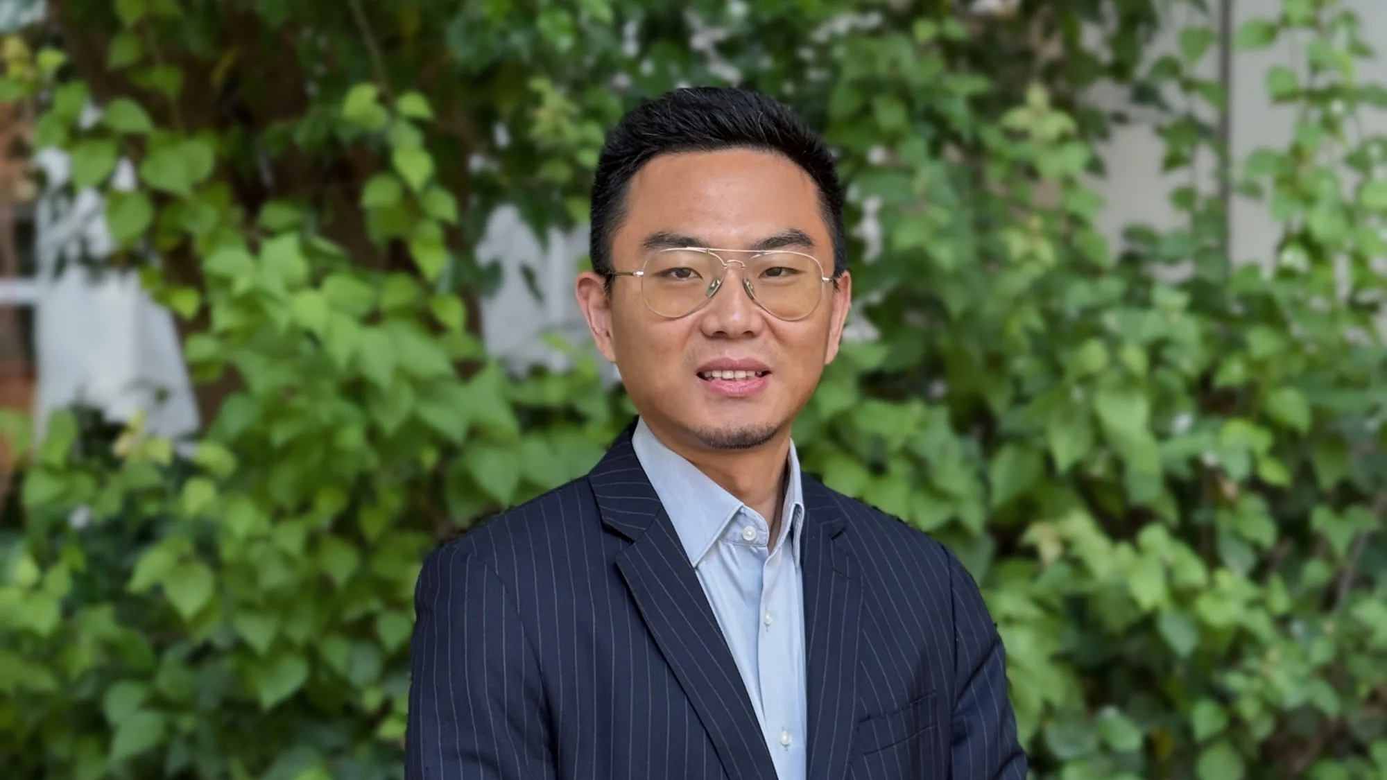 Kevin in a light blue shirt and dark suit in front of a leafy green border