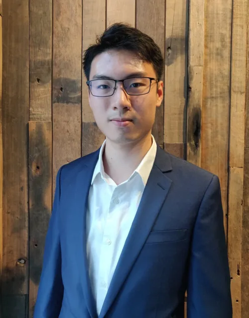 Vincent Kong stood in front of a wooden wall in a white open collar shirt and blue jacket