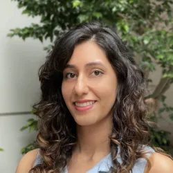 A person with shoulder‑length curly hair stands outdoors in front of a light‑coloured wall and green foliage. The individual is wearing a sleeveless top, and trees and plants fill the background.