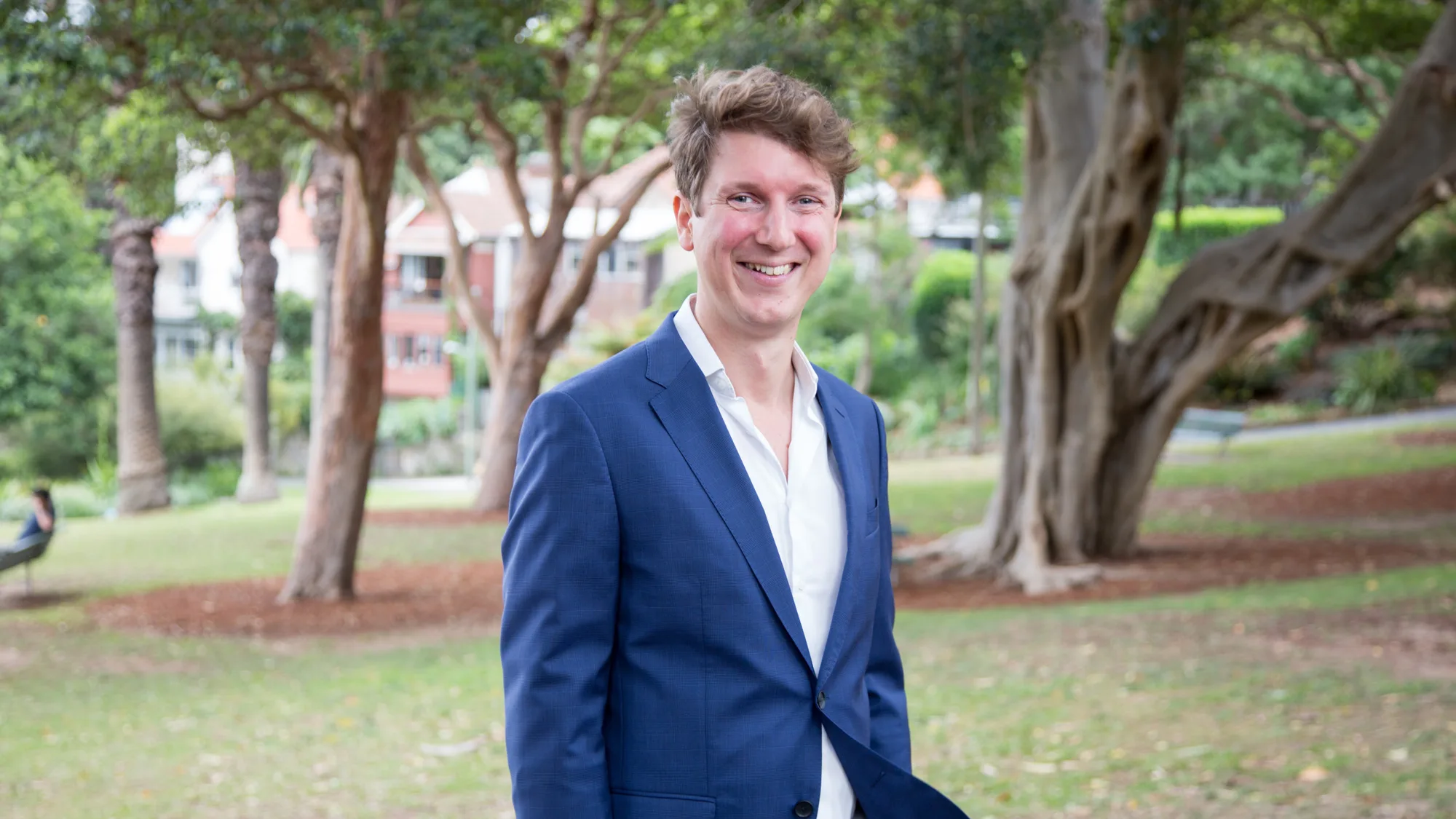 Alan wearing a white open collar shirt and blue suit in a suburban park