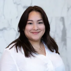 A person with long dark hair wearing a white shirt and a small necklace stands in front of a light gray marble wall. The image shows the upper body and shoulders against the textured stone background.