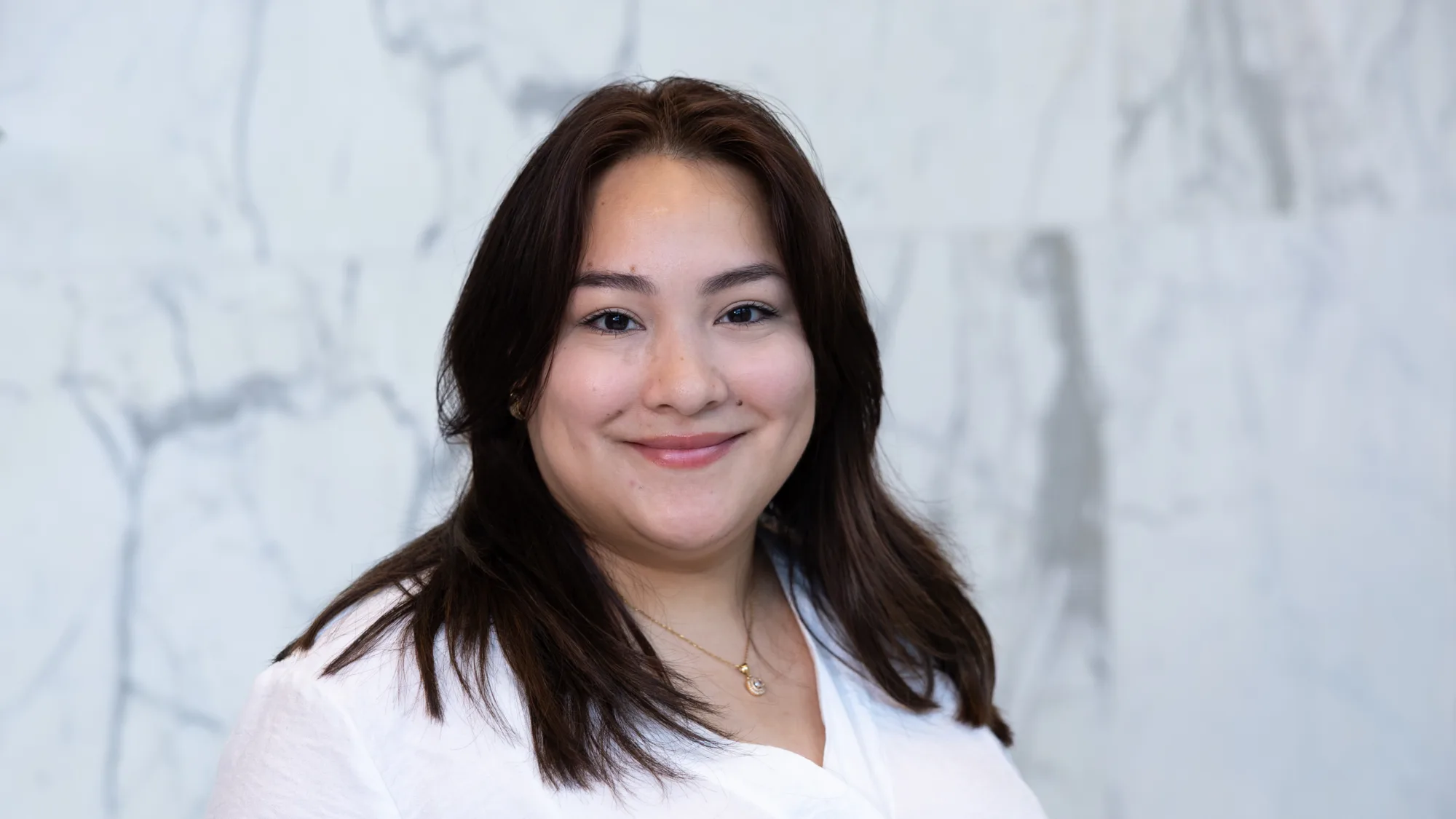A person with long dark hair wearing a white shirt and a small necklace stands in front of a light gray marble wall. The image shows the upper body and shoulders against the textured stone background.
