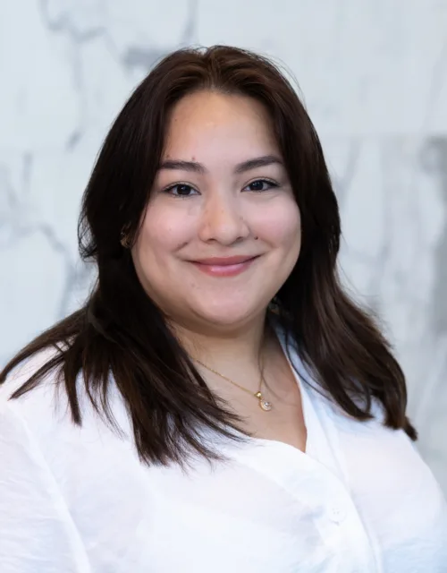 A person with long dark hair wearing a white shirt and a small necklace stands in front of a light gray marble wall. The image shows the upper body and shoulders against the textured stone background.