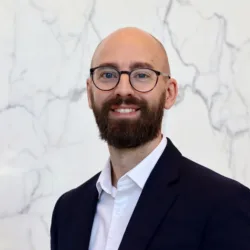 Andy in a dark suit jacket and white dress shirt stands in front of a white marble wall with grey veining.