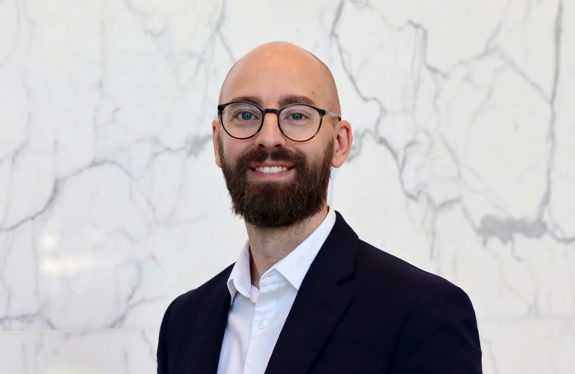 Andy in a dark suit jacket and white dress shirt stands in front of a white marble wall with grey veining.