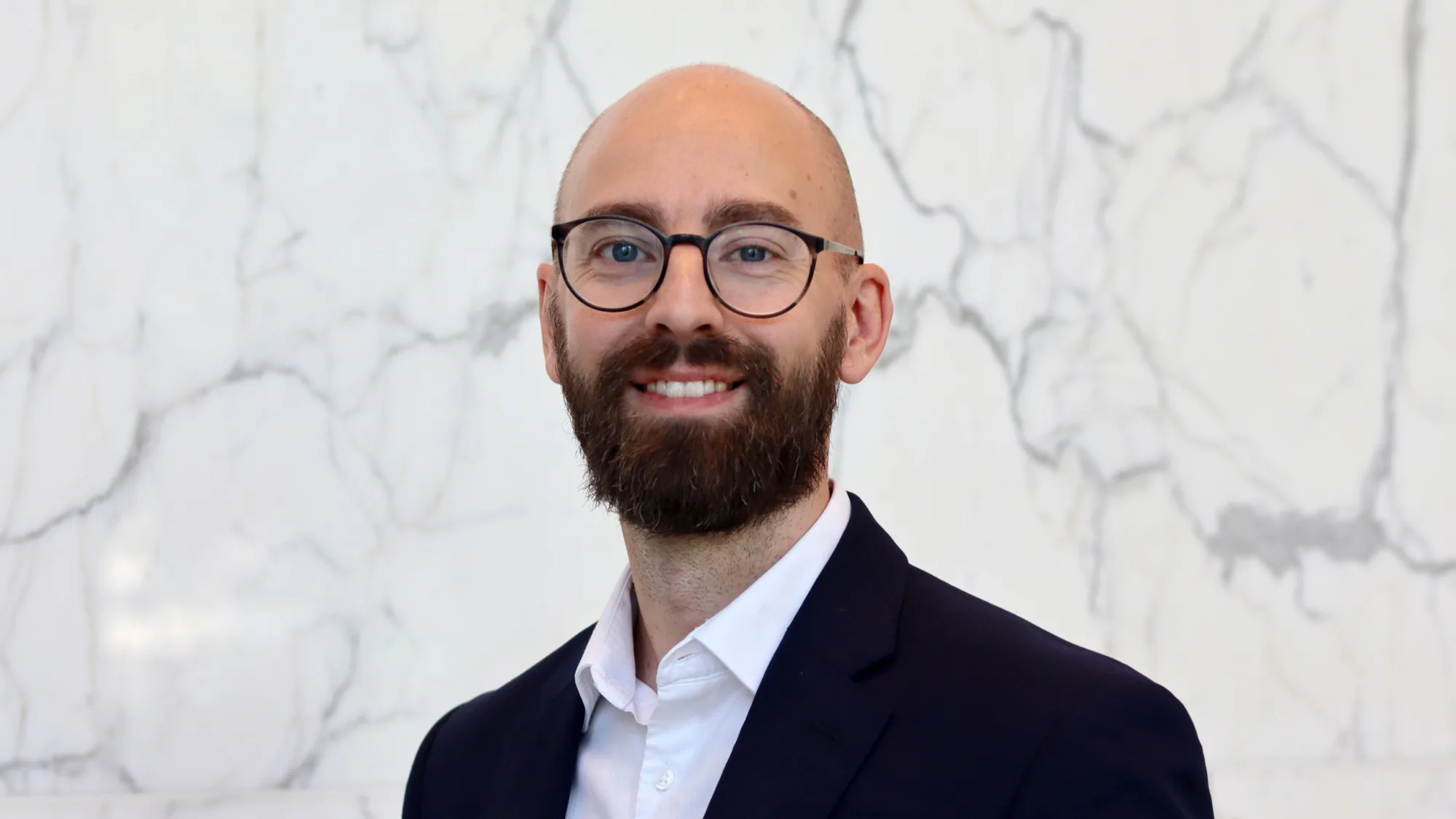 Andy in a dark suit jacket and white dress shirt stands in front of a white marble wall with grey veining.