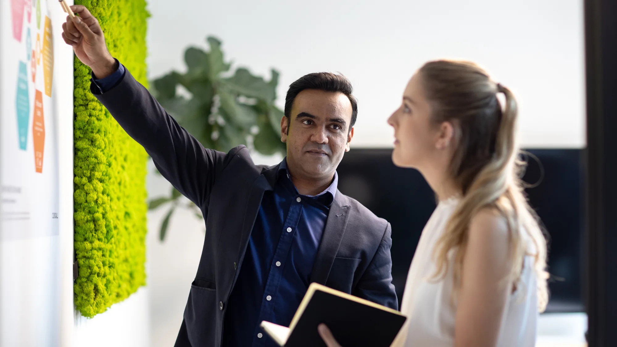 Chadik with a colleague pointing to a wall mounted infographic next to a moss wall panel