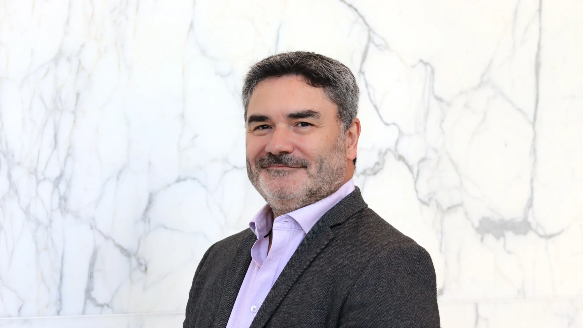 Head and shoulders shot of Drew against a white marble background wearing a pale pink shirt and grey blazer