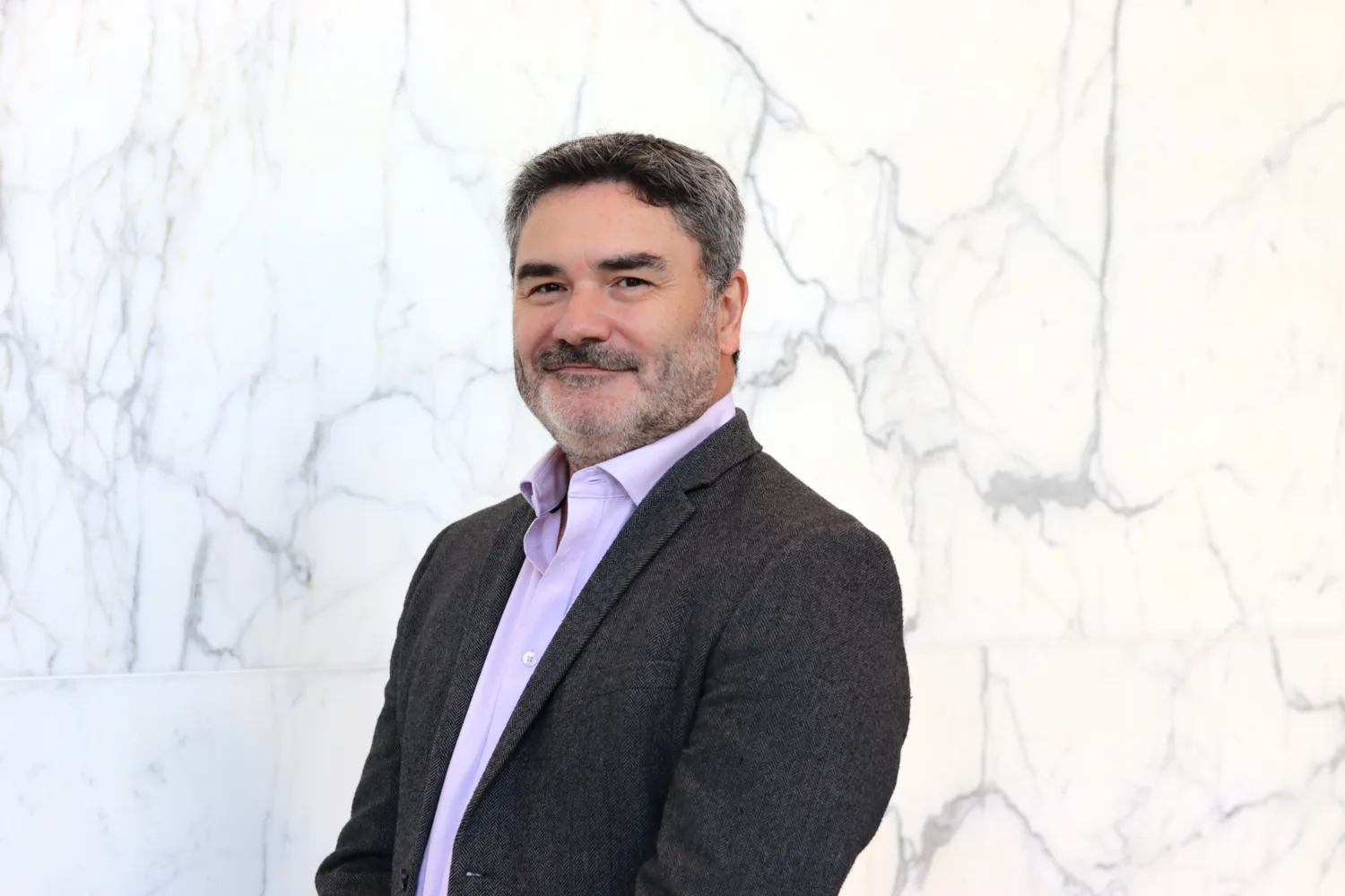 Head and sholders shot of Drew against a white marble background wearing a pale pink shirt and grey blazer