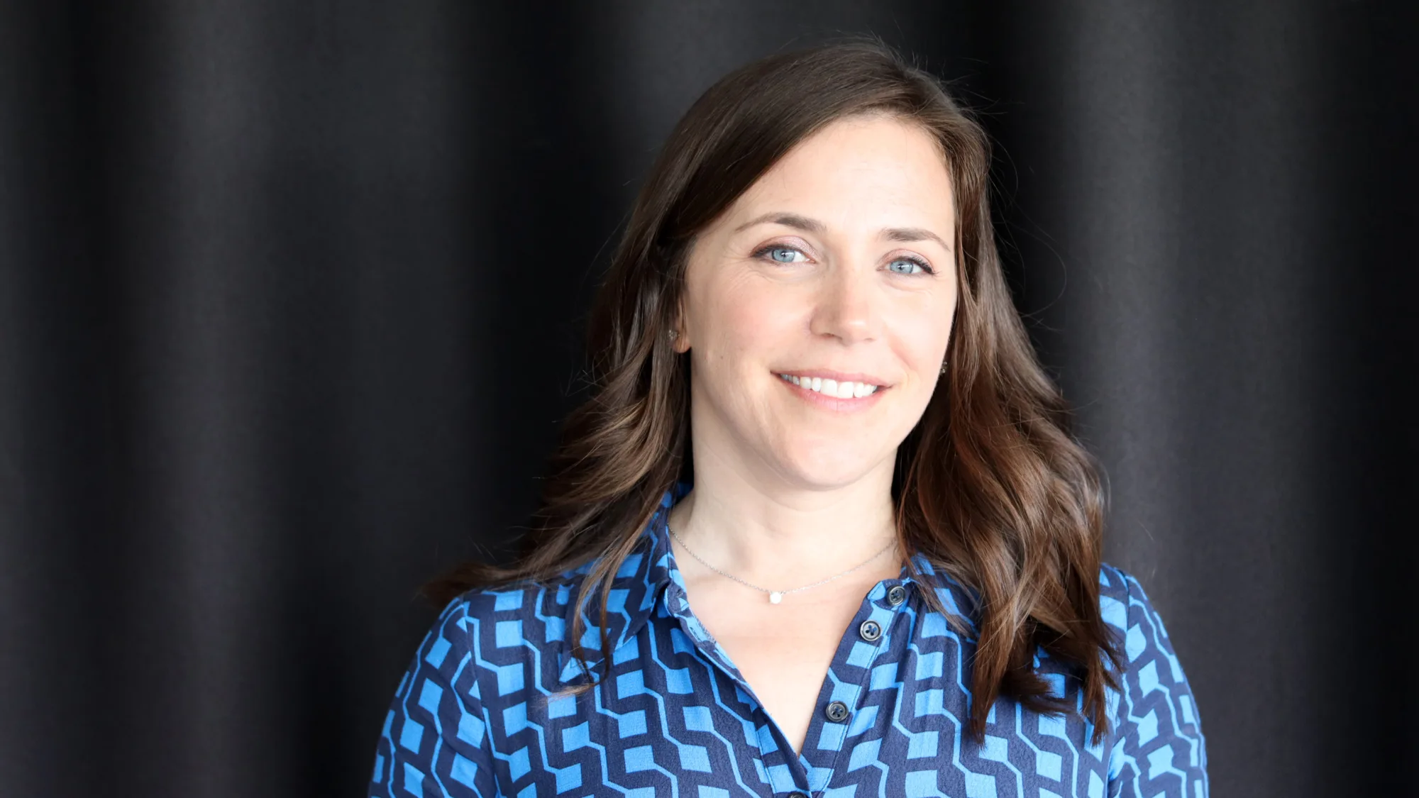 Frances in a blue buttoned top in front of a dark fabric wall