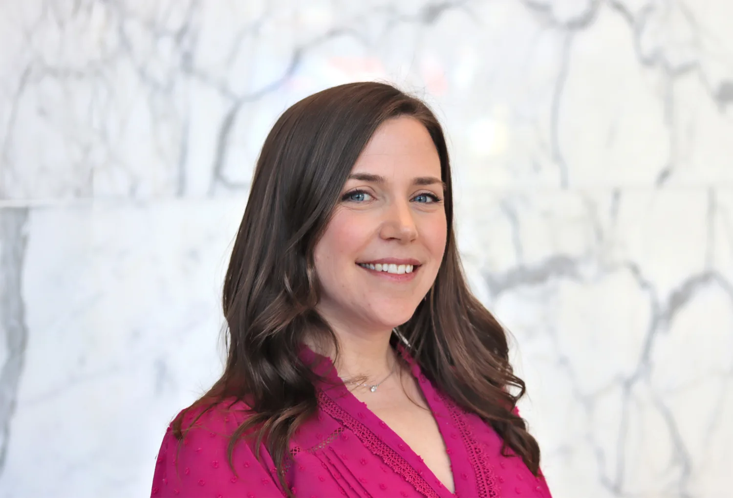 A person with long wavy brown hair is shown from the shoulders up, wearing a bright pink textured top and a small necklace. They stand in front of a light grey marble wall with dark veining.