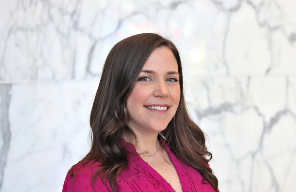 A person with long wavy brown hair is shown from the shoulders up, wearing a bright pink textured top and a small necklace. They stand in front of a light grey marble wall with dark veining.
