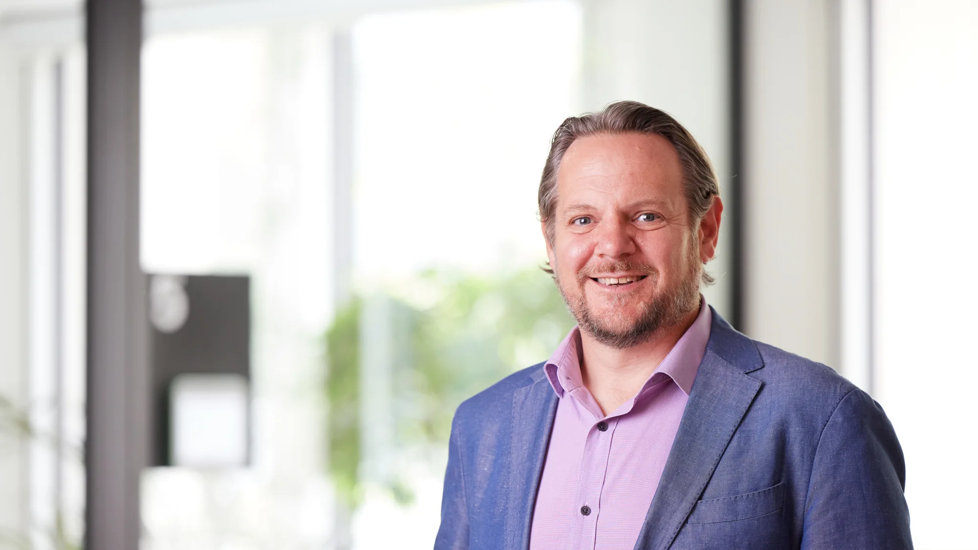 Head and sholders shot of Grant wearing a pink shirt and a blue blazer against the london office background