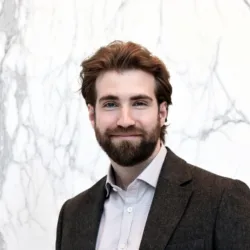 Jordan in an open collar shirt and dark suit jacket smiling to camera in front of a grey marbled wall