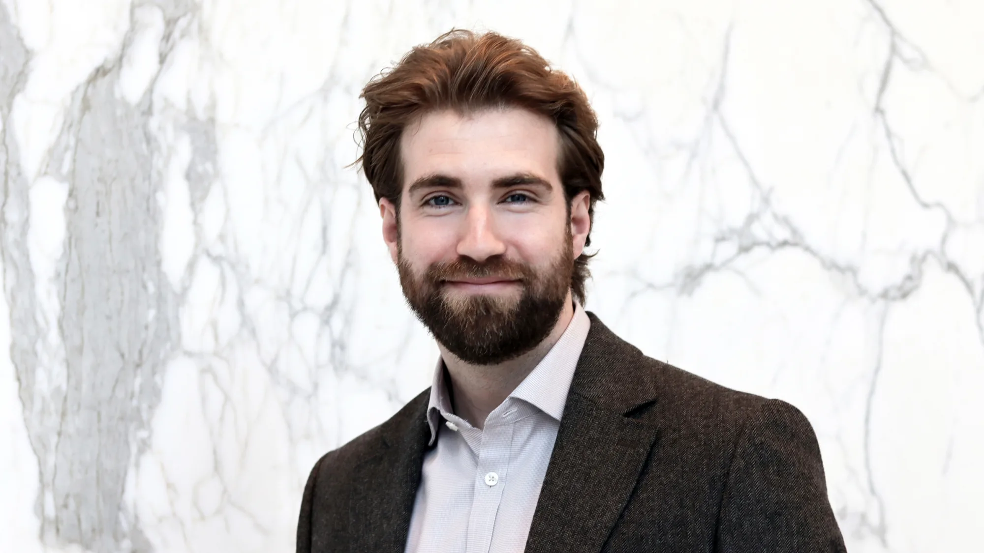 Jordan in an open collar shirt and dark suit jacket smiling to camera in front of a grey marbled wall