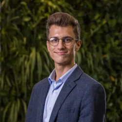 Laszlo in a pale blue open collar shirt and suit jacket in front of a green living wall smiling to camera