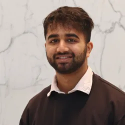 A person wearing a dark sweatshirt over a white collared shirt stands in front of a light marble-patterned wall.