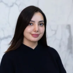 A person with straight dark hair wearing a dark V‑neck sweater is shown from the shoulders up, standing against a light grey marble wall with visible veining patterns in the background.