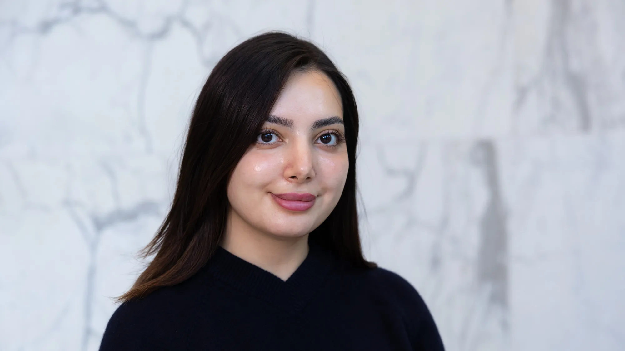 A person with straight dark hair wearing a dark V‑neck sweater is shown from the shoulders up, standing against a light grey marble wall with visible veining patterns in the background.