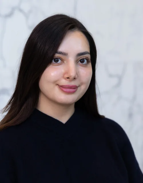 A person with straight dark hair wearing a dark V‑neck sweater is shown from the shoulders up, standing against a light grey marble wall with visible veining patterns in the background.