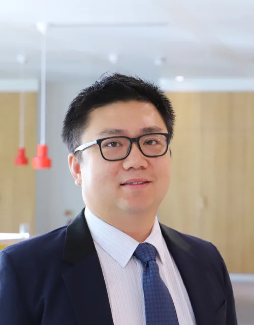 Qian Li standing in a bright room with a row of cabinet doors and hanging red lights slightly out of focus in the background